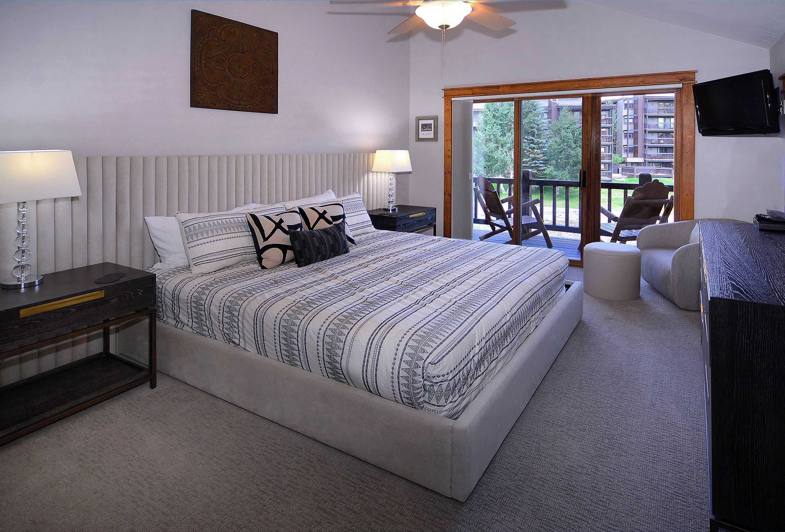 A bedroom with a large bed, a nightstand, a desk, a chair, and a balcony with a view of buildings.