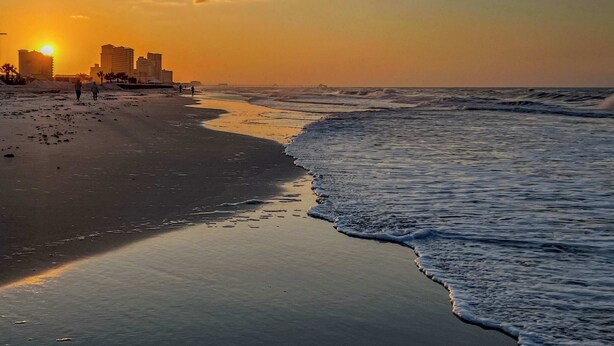Een strand met een zonsondergang, golven en gebouwen op de achtergrond.