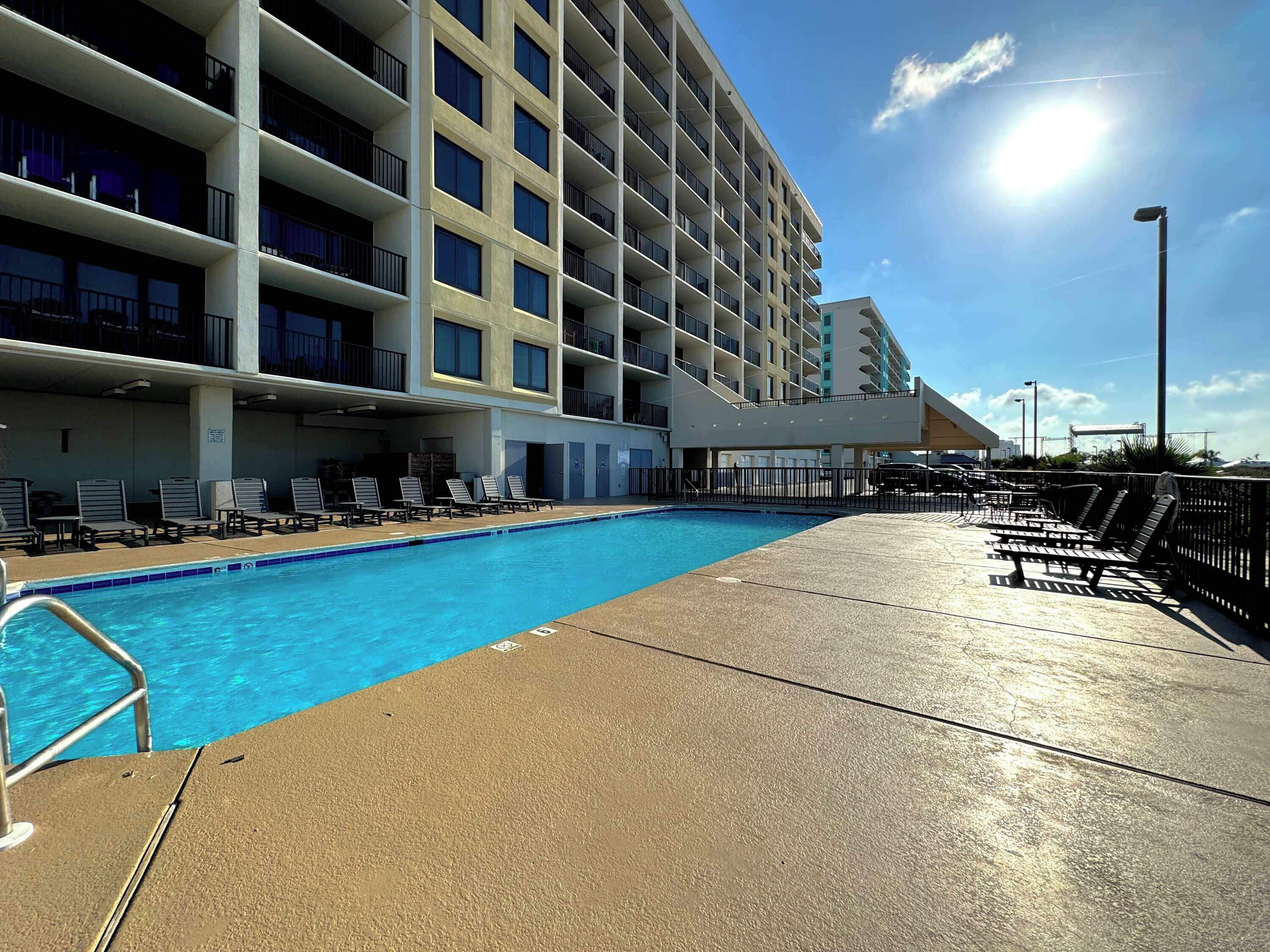 Un espace piscine d’hôtel avec une piscine à l’eau d’un bleu limpide, des chaises longues et un bâtiment doté de nombreux balcons.