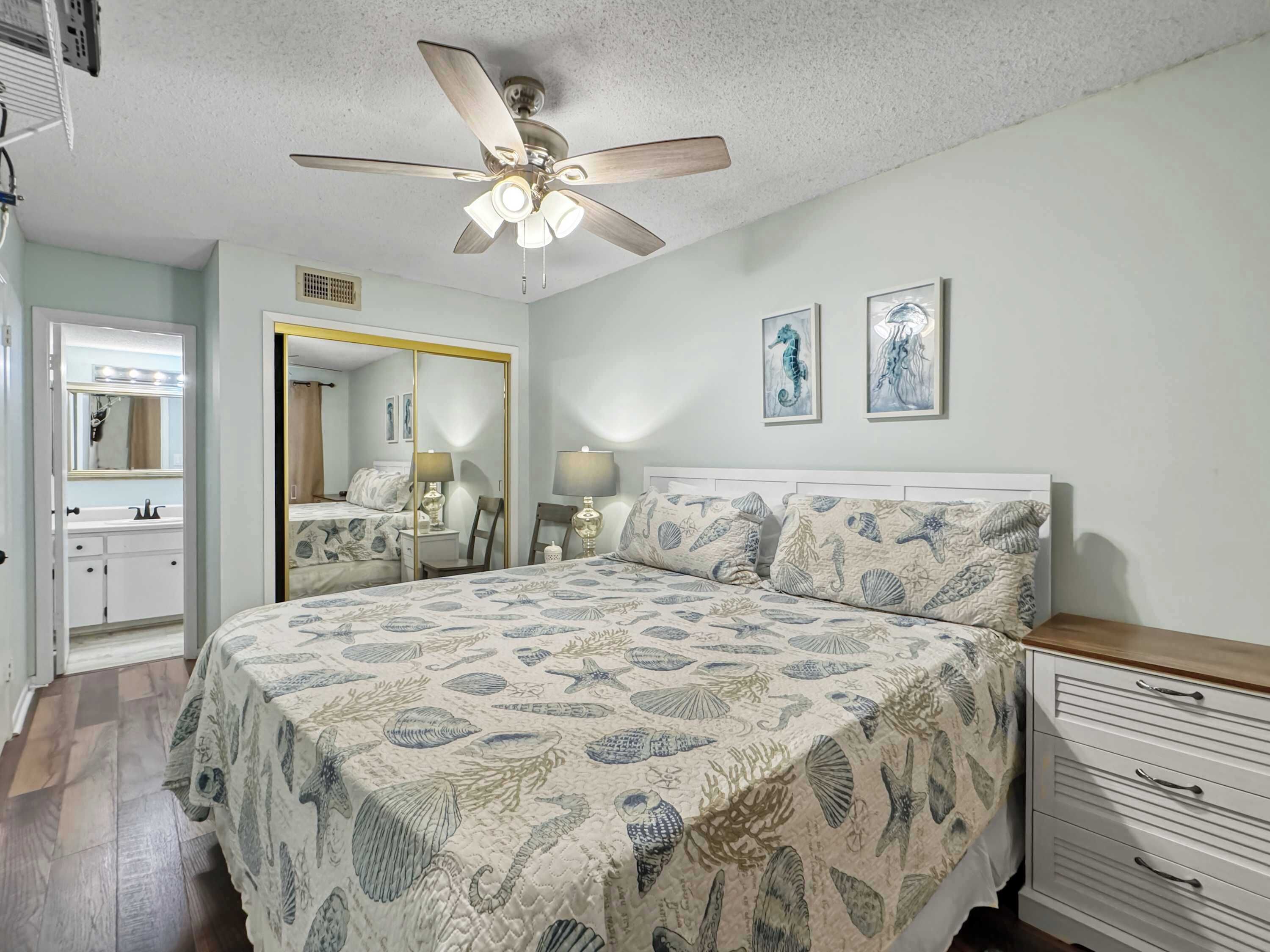 A bedroom with a large bed, a ceiling fan, a nightstand, a dresser, a mirror, and two framed seahorse paintings on the wall.