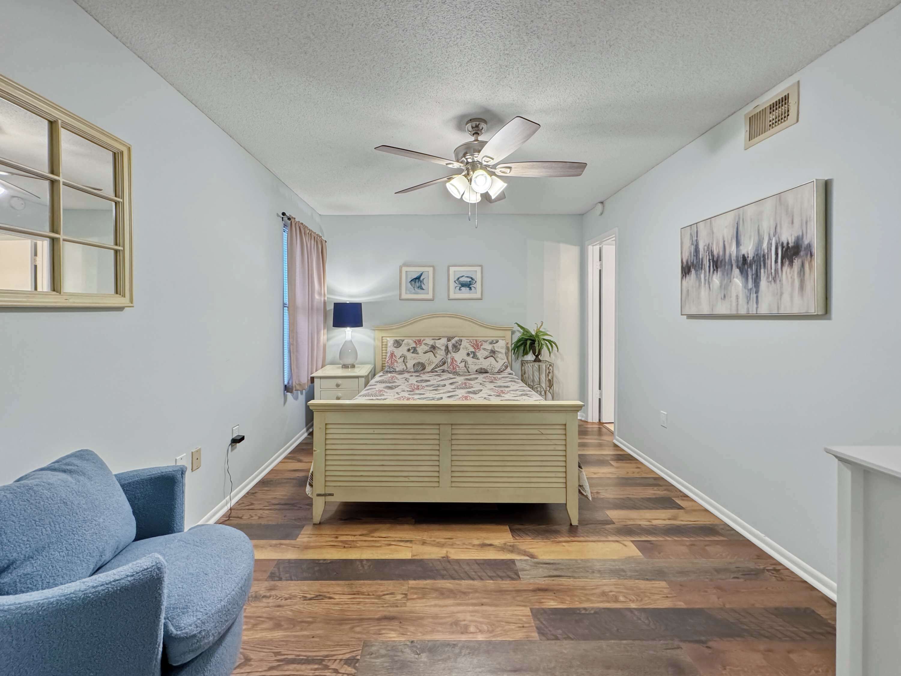 A bedroom with a bed, a ceiling fan, a window with curtains, a bedside table with a lamp, a blue chair, and a painting on the wall.