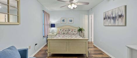 A bedroom with a bed, a ceiling fan, a window with curtains, a bedside table with a lamp, a blue chair, and a painting on the wall.