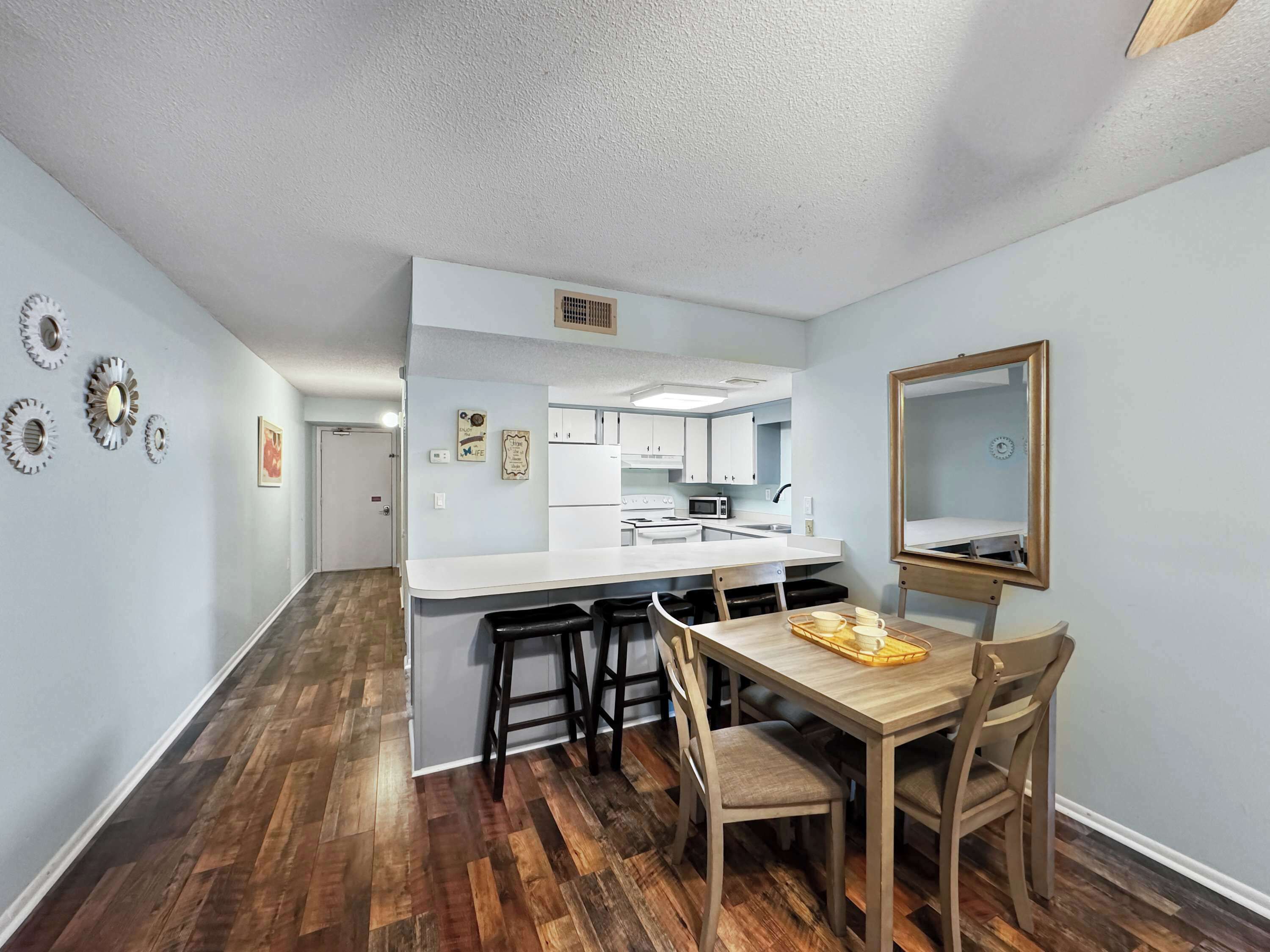 A compact kitchen with white cabinets, a microwave, and a sink. There is a dining area with a wooden table and chairs, and a mirror on the wall.