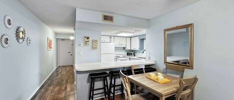 A compact kitchen with white cabinets, a microwave, and a sink. There is a dining area with a wooden table and chairs, and a mirror on the wall.