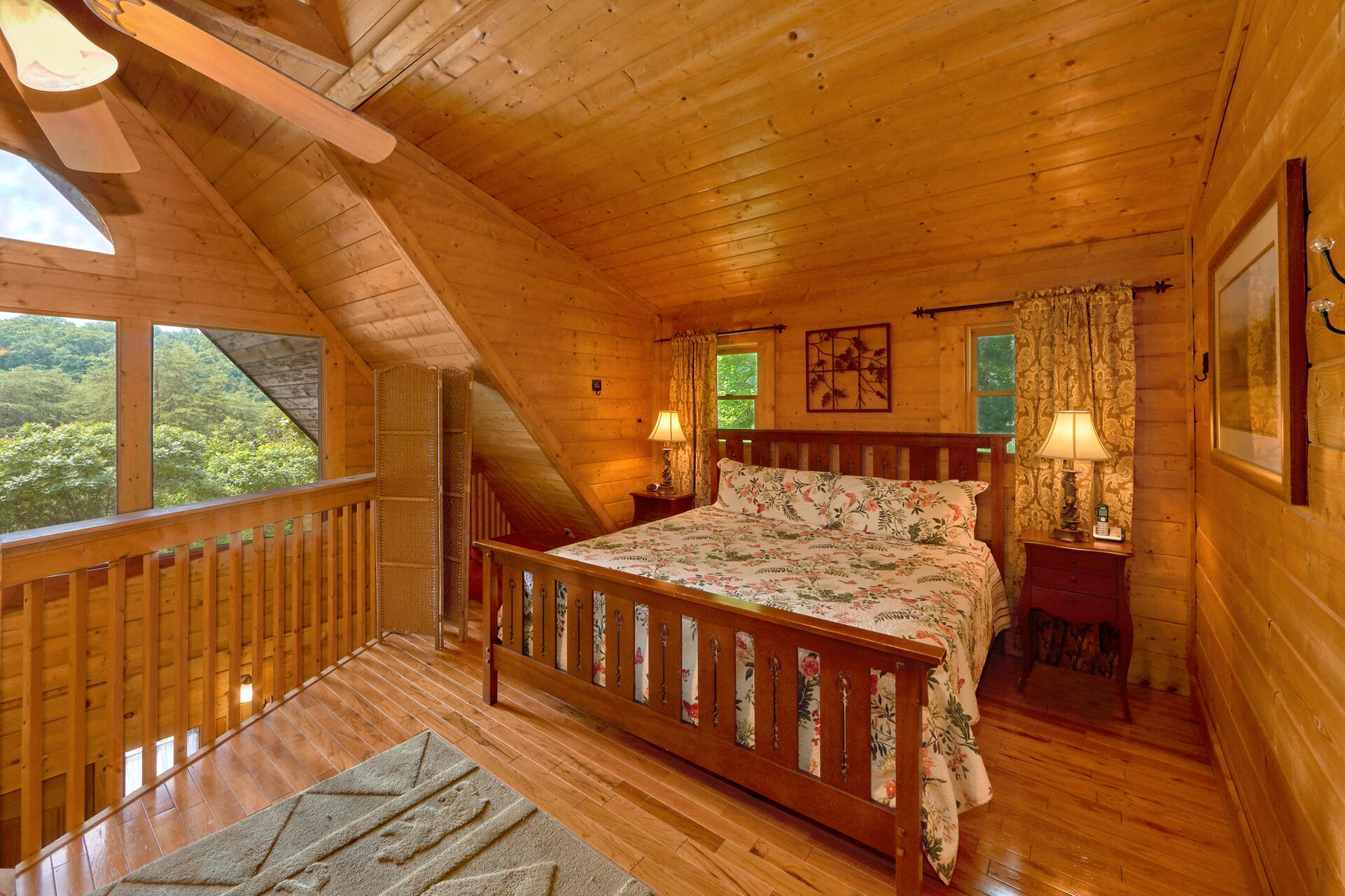 A wooden cabin bedroom with a bed, two bedside lamps, a window, and a view of greenery outside.