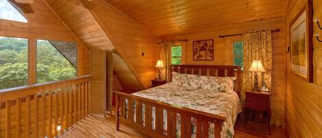 A wooden cabin bedroom with a bed, two bedside lamps, a window, and a view of greenery outside.