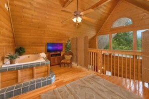 A wooden cabin interior with a wooden deck, a bathtub, a television, and a ceiling fan.