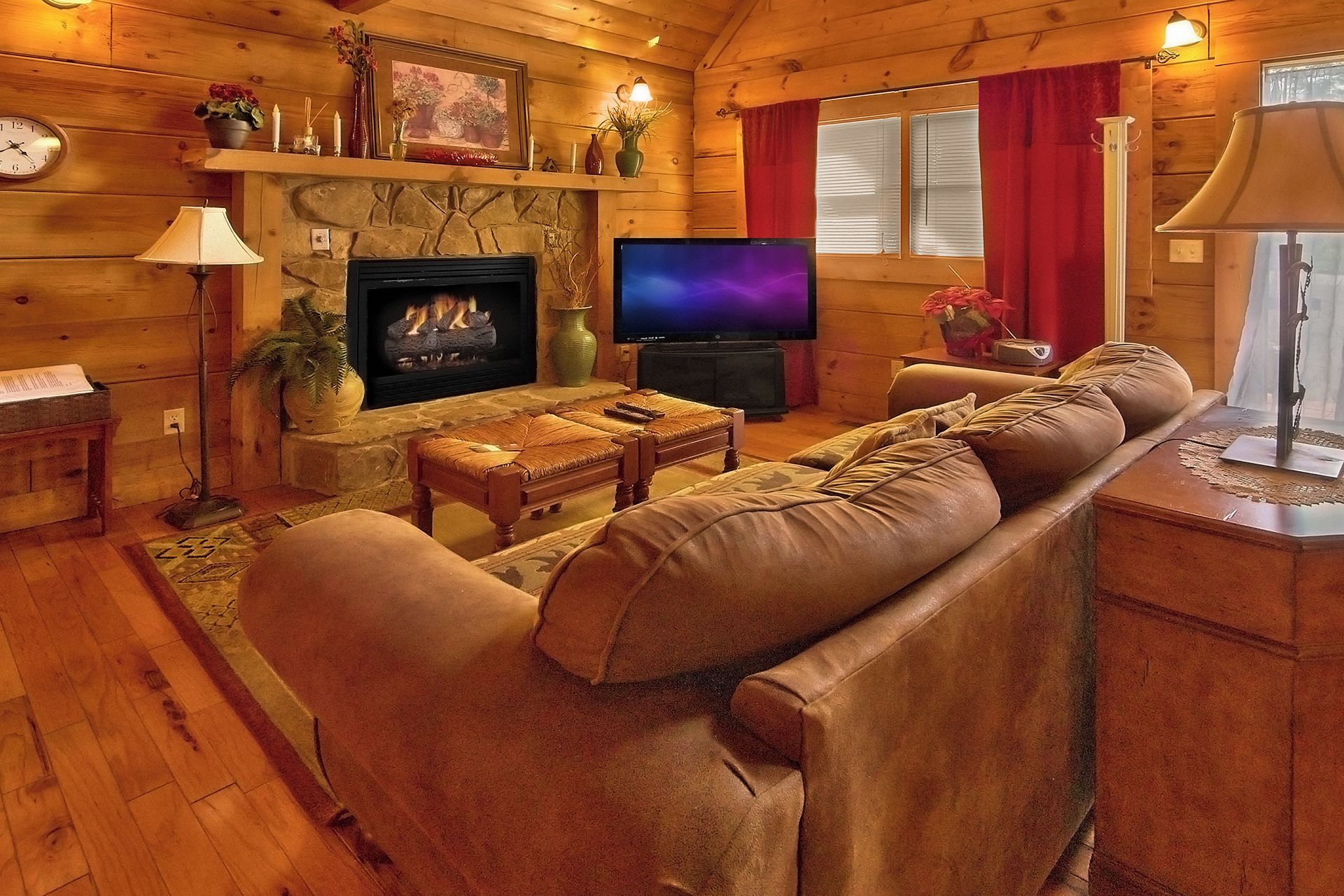 Een gezellig blokhutinterieur met een stenen open haard, een televisie, leren banken en houten wanden.
