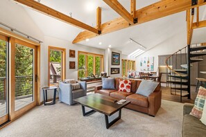 A spacious living area with a wooden ceiling, large windows, a fireplace, and a staircase.