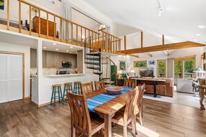 A spacious living area with a wooden staircase, a dining table with chairs, and a kitchen area.