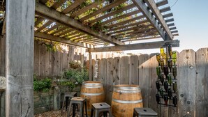 Eine Holzhütte mit Weinregal, zwei Fässern und Hockern in einem Garten.
