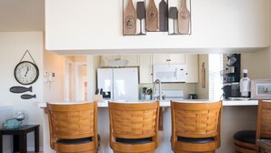 Eine Küche mit weißen Schränken, einem Kühlschrank, einer Mikrowelle und einem Waschbecken. Drei Holzstühle mit gewebten Rücken sind vorhanden. Eine Uhr hängt an der Wand, daneben ein Fischdekor.