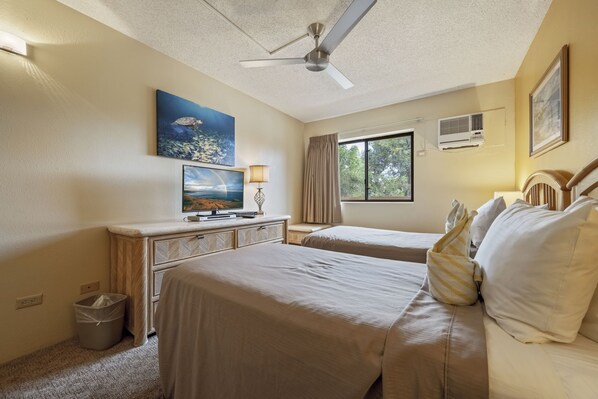 Una habitación de hotel con dos camas, un ventilador de techo, una ventana con cortinas, un equipo de aire acondicionado y obras de arte en las paredes.