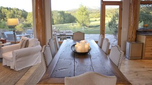 Un espace repas comprenant une table en bois, des chaises à revêtement blanc et une vue sur un terrain de golf grâce à de grandes fenêtres.