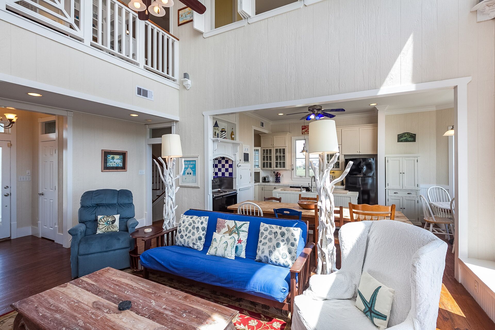 A spacious living room with a blue sofa, a wooden coffee table, and a dining area with a wooden table and chairs. A kitchen with white cabinets and a black refrigerator is visible in the background.