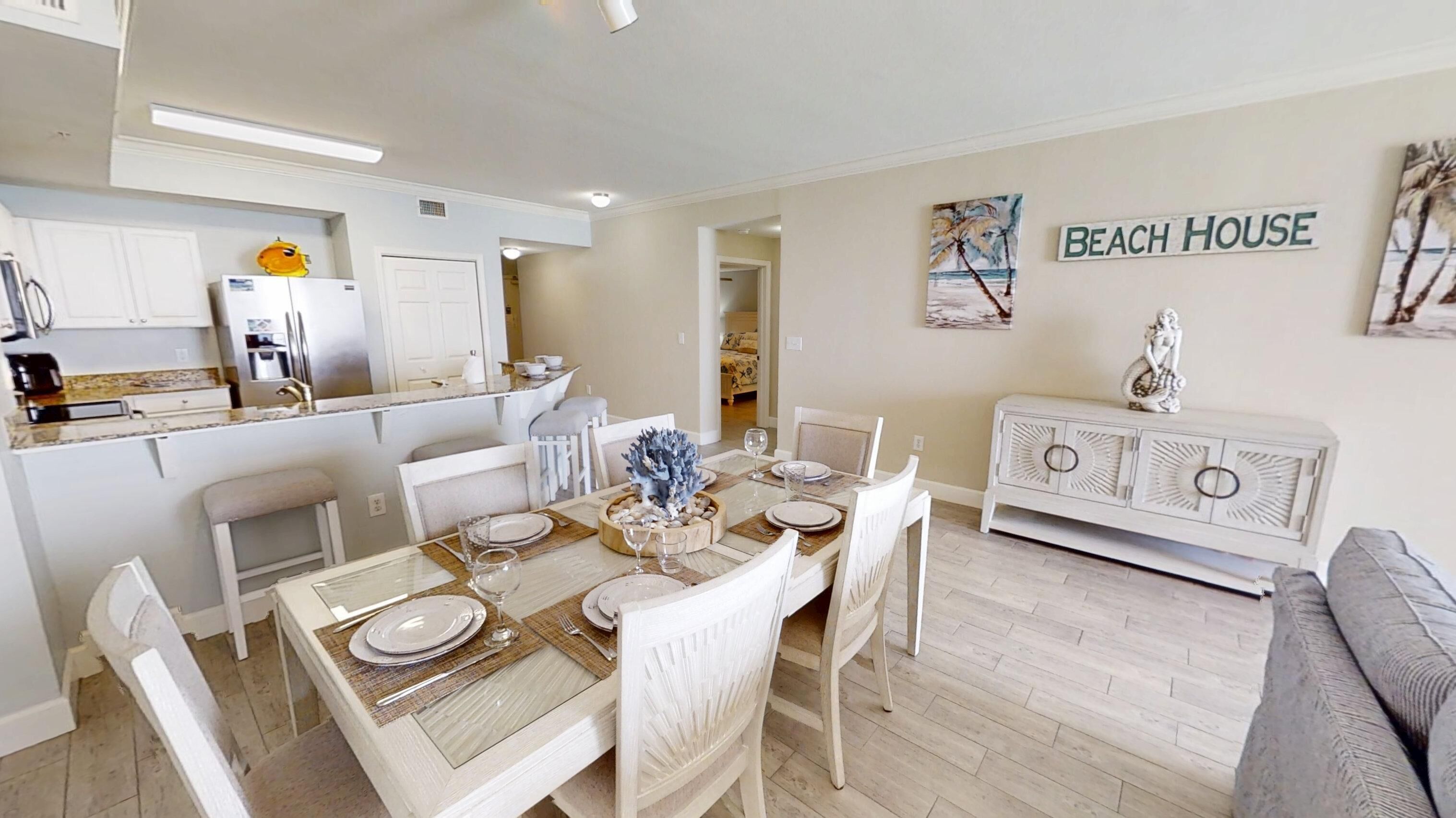 A dining area with a table set for four, white chairs, a white cabinet, and a wall sign that reads "BEACH HOUSE".