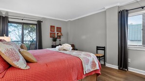 Een slaapkamer met een groot bed, een stoel, een klein tafeltje met een lamp en een raam met gordijnen.