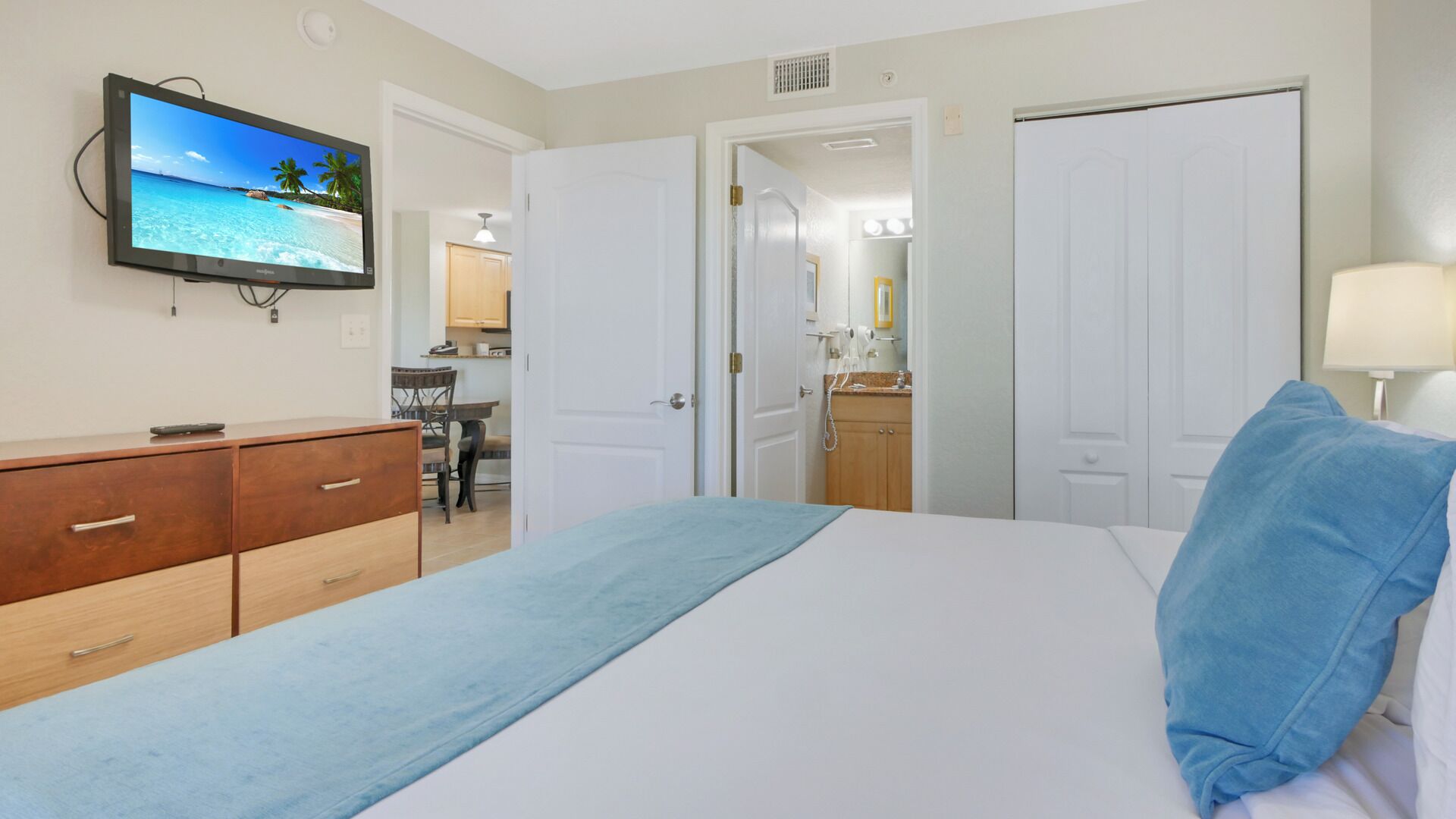 Habitación de hotel con cama, televisor mostrando una playa, escritorio con silla y baño visible a través de una puerta abierta.