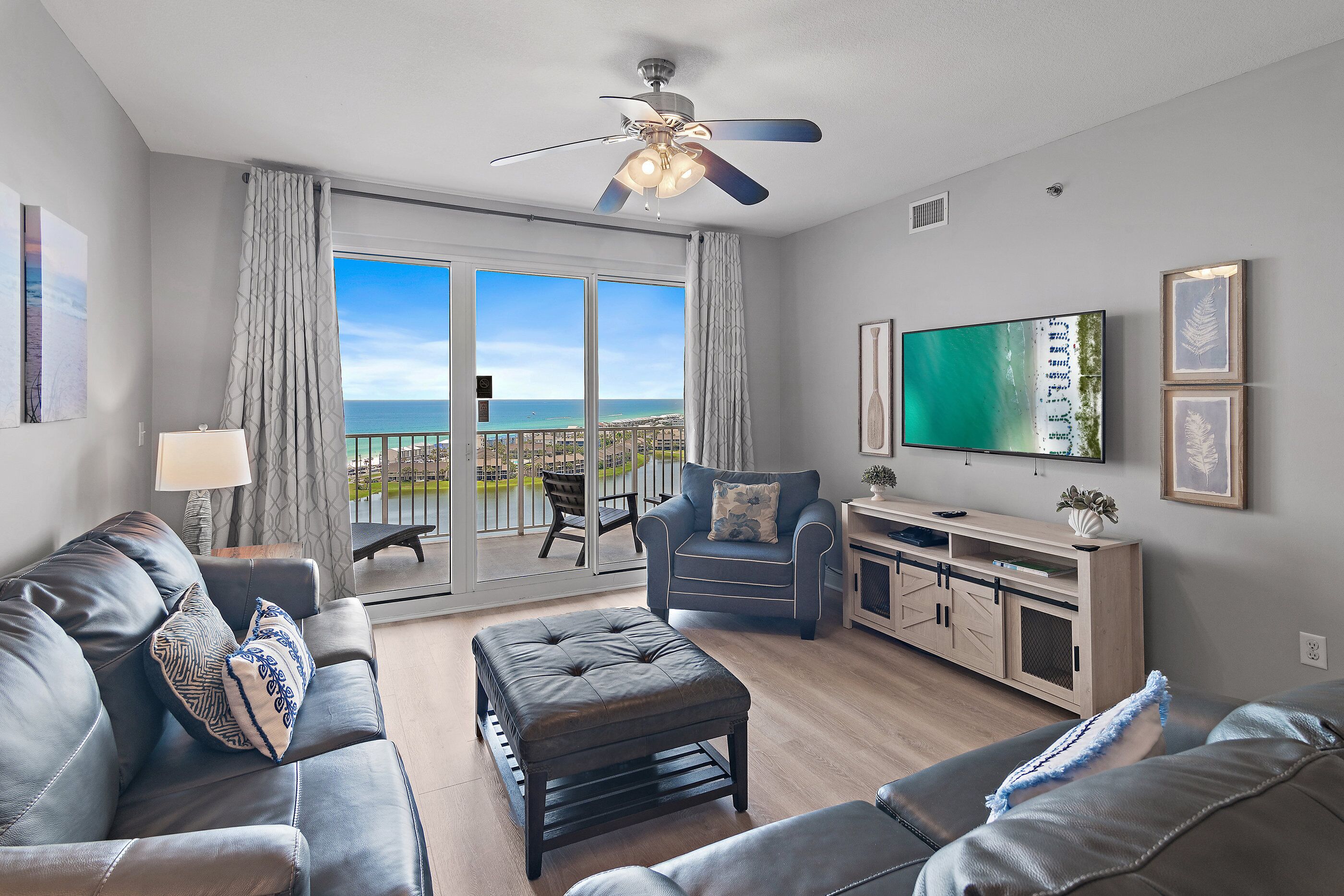 A modern living room with a leather sofa, ottoman, and armchair, a flat-screen TV, and a balcony with a view of the ocean.