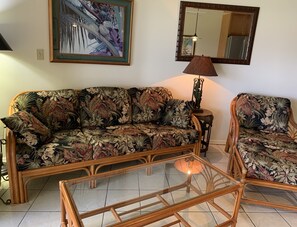 A living room with a floral sofa, a glass coffee table, a side table with a lamp, and a framed painting on the wall.