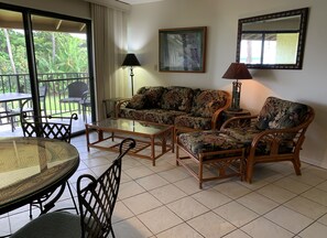 A living room with a glass coffee table, a sofa, two armchairs, a dining table with chairs, a lamp, and a painting on the wall.