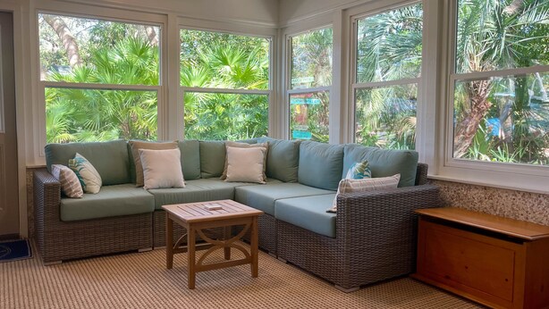 A cozy room with a sectional sofa, a small wooden table, and large windows offering a view of lush greenery.