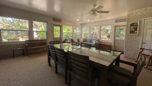 A spacious dining area with a large table and chairs, surrounded by large windows offering views of greenery.