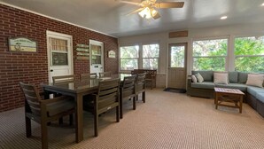 A room with a brick wall, a dining table with chairs, a sofa, and large windows with a view of greenery.