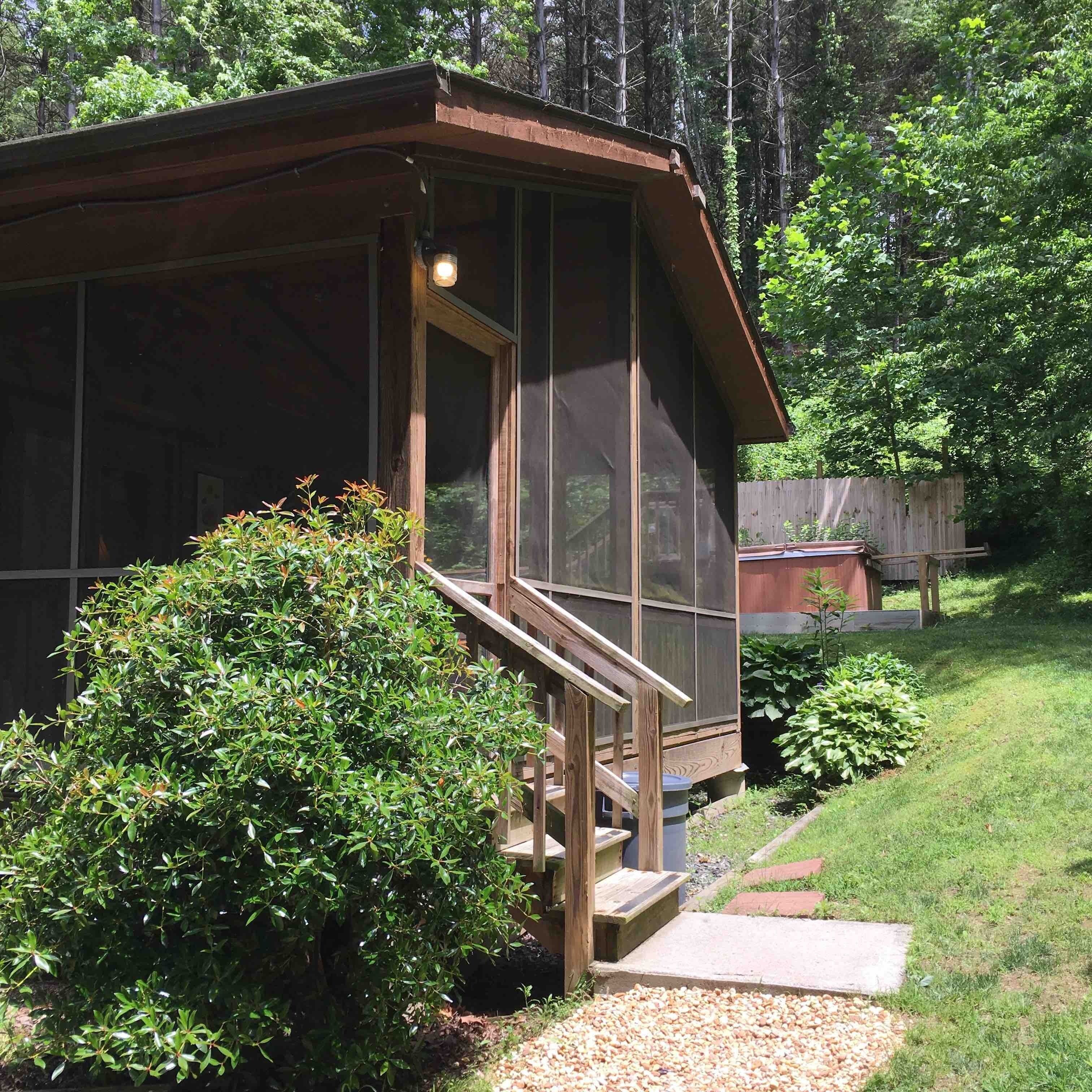 A small wooden cabin with a porch and a large window, surrounded by lush greenery.