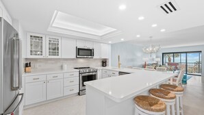 A modern kitchen with white cabinetry, a central island with bar stools, stainless steel appliances, and a large window overlooking a scenic view. - Sanddollar #A202 is a 3/2 beachfront Vacation Rental on Sanibel Island (Sanibel)