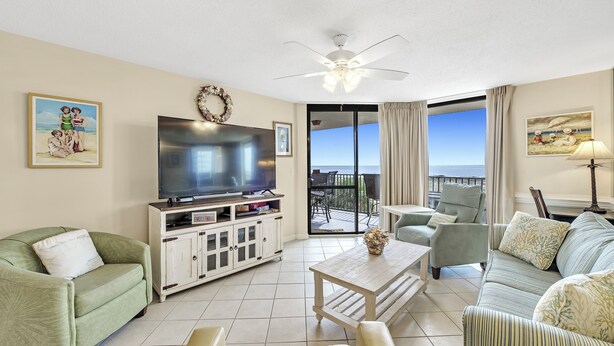 A living room with a flat-screen TV, a ceiling fan, a sofa, a coffee table, and a view of the ocean through large windows. - Phoenix VII 213 | Beachfront | Pools | Fitness Center | My Beach Getaways (Orange Beach)
