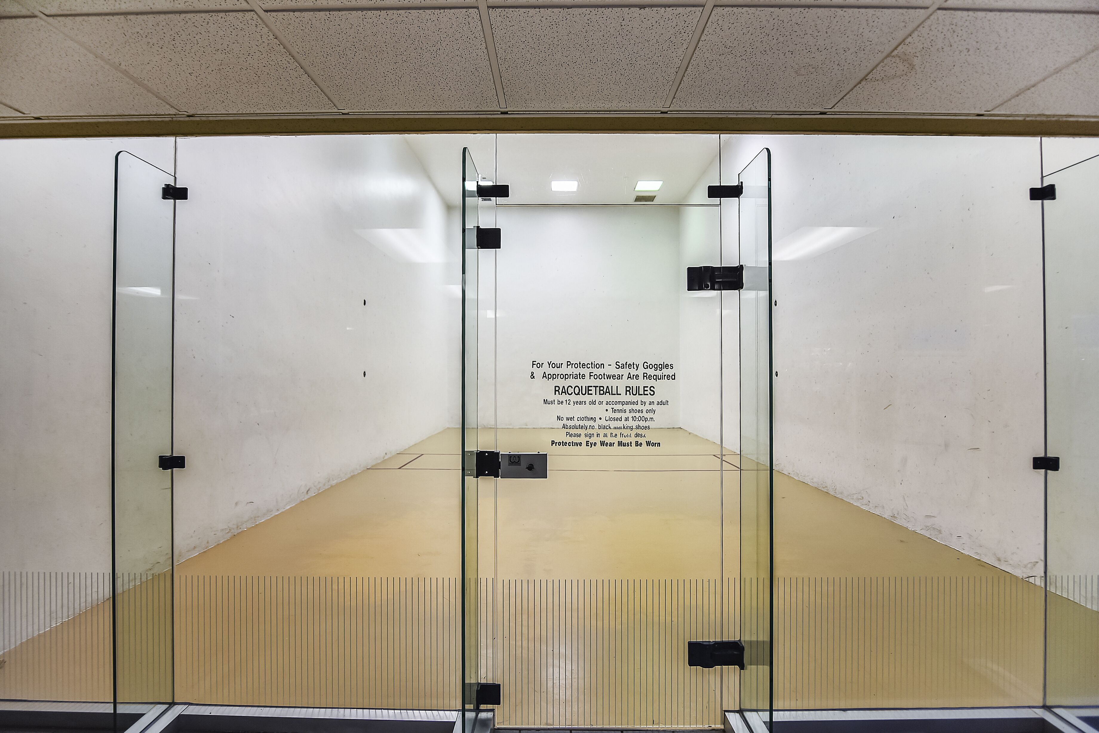 A racquetball court with glass walls and a wooden floor, featuring rules and safety instructions.