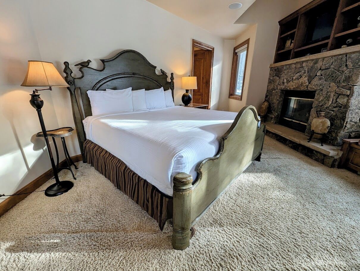 A bedroom with a large bed, two bedside lamps, a stone fireplace, and a window.