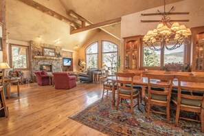 A spacious living room with wooden floors, a stone fireplace, and large windows offering views of the outdoors.