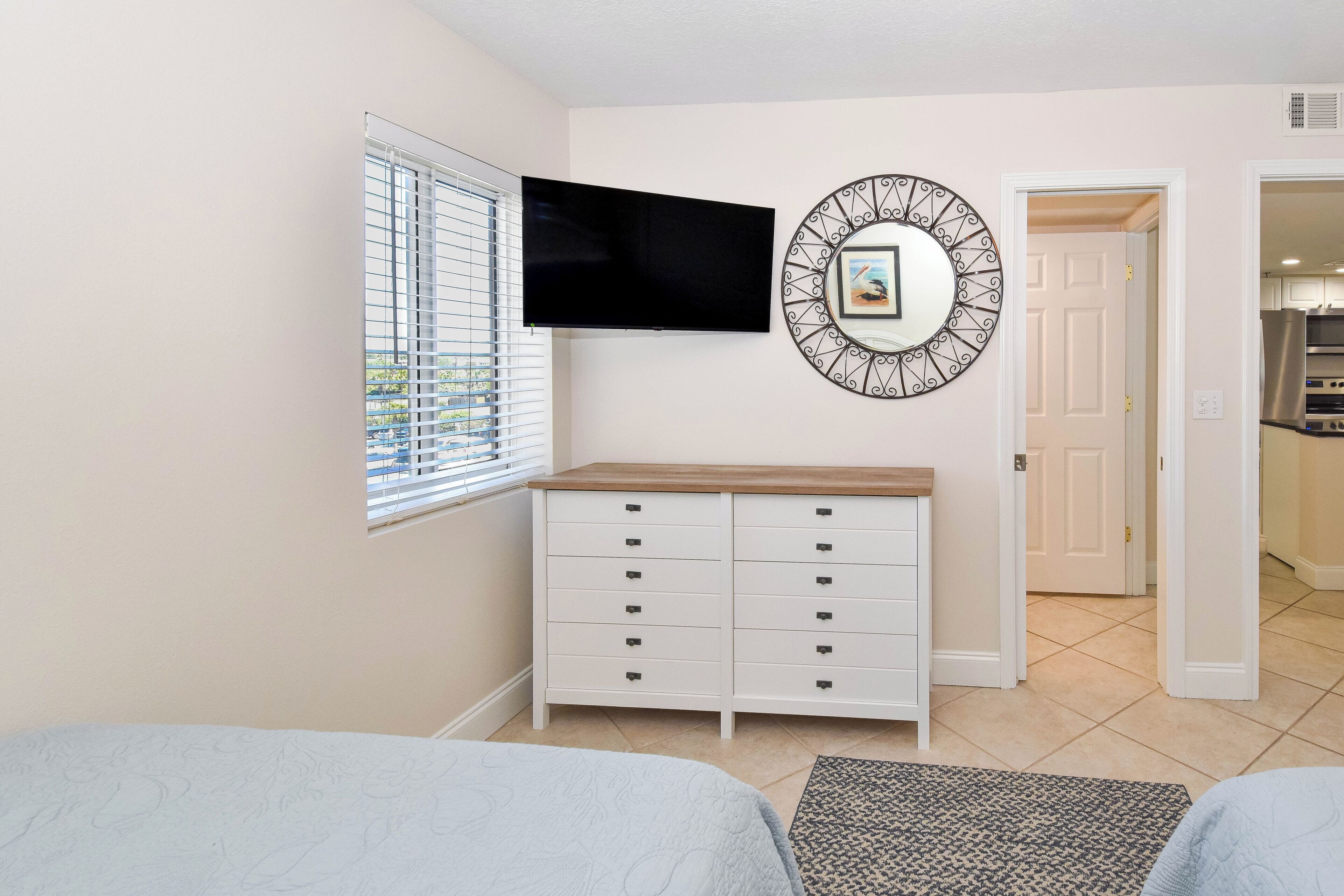 A bedroom with a TV, a dresser, a window with blinds, and a door leading to a kitchen.