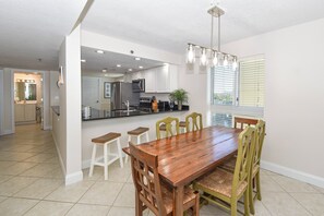 A modern kitchen with a dining area, white cabinets, a stainless steel refrigerator, and a wooden dining table with chairs. - Corner condo at Destin Towers overlooking Gulf-front pool & beach! (Destin)