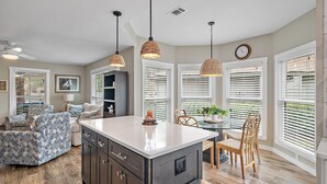 A modern kitchen with a central island, pendant lighting, and large windows with blinds.