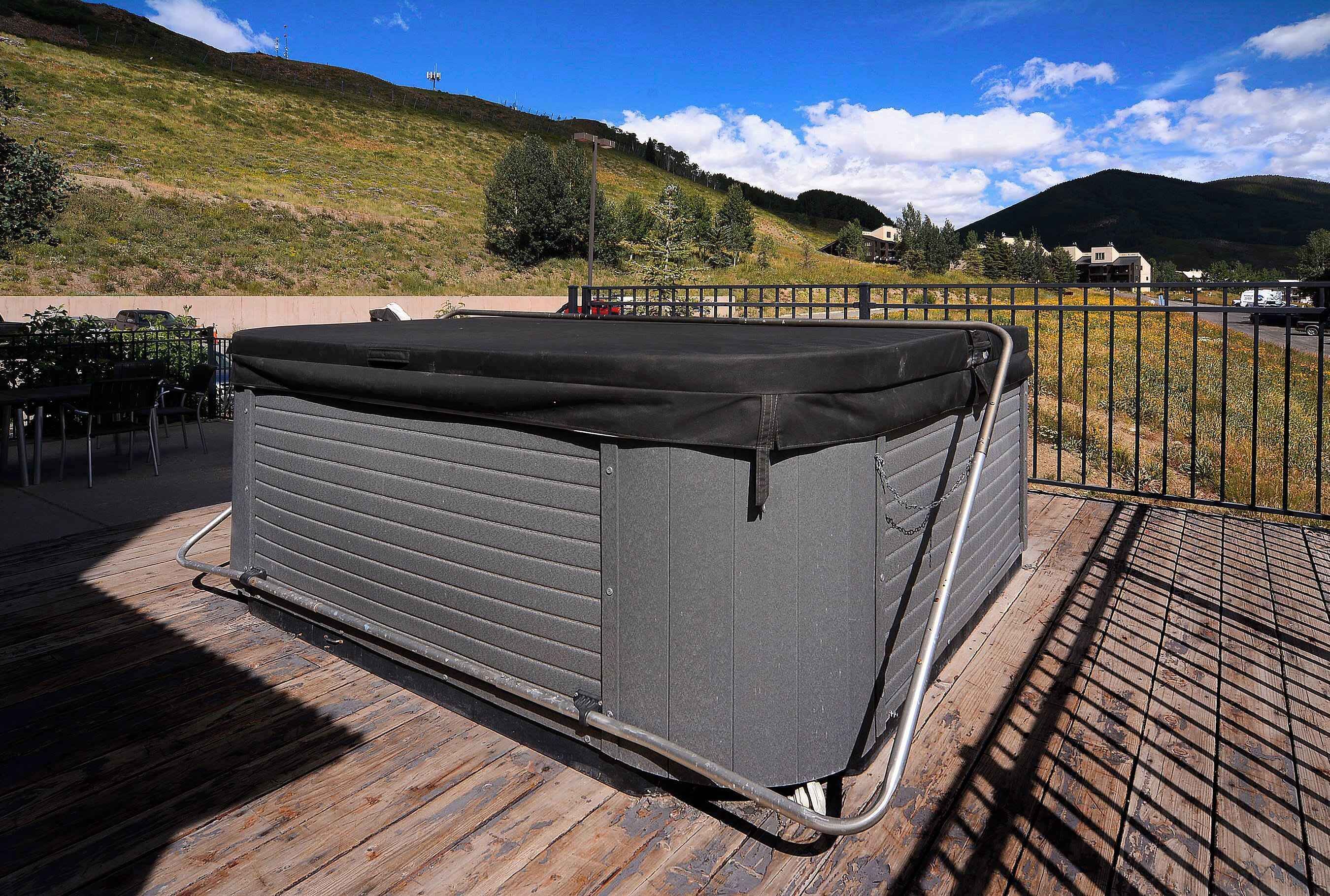 Una vasca idromassaggio su un pontile di legno con ringhiera metallica, con vista su un paesaggio collinare con case.
