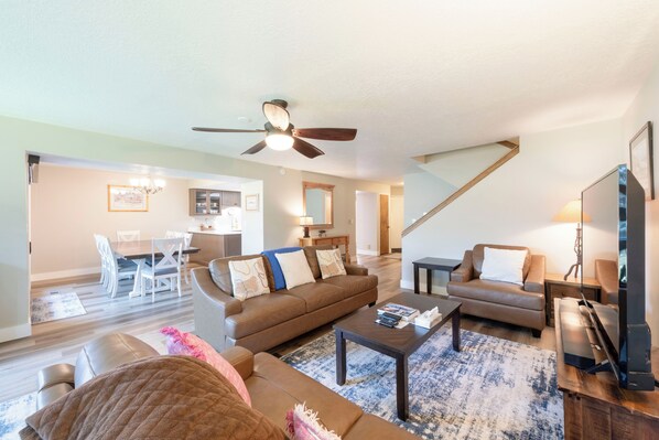A spacious living room with a brown sofa, a coffee table, and a television. There is a dining area with a table and chairs in the background.