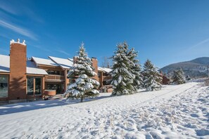 A snow-covered residential area with houses, a clear blue sky, and a hilly landscape in the background.