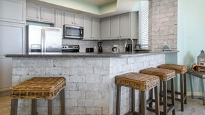 Une cuisine avec des meubles blancs, un pare-feu en pierre, un réfrigérateur en acier inoxydable, une machine à café et trois tabourets en osier.