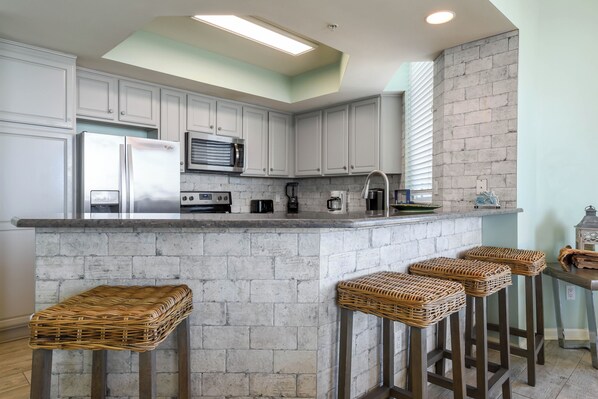 A kitchen with white cabinets, a stone backsplash, a stainless steel refrigerator, a coffee maker, and three wicker stools. - Gulf-front Beachy Condo w/ Easy Beach Access & Large Balcony in Destin! 🏖🌊☀ (Destin)