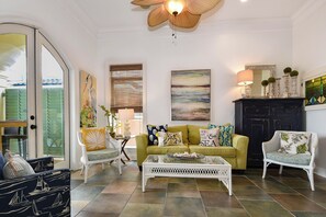 A living room with a green sofa, white armchairs, a glass coffee table, and a ceiling fan.