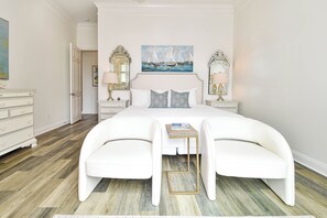 A bedroom with a large bed, two white chairs, a nightstand with lamps, and a painting of a sailboat on the wall.