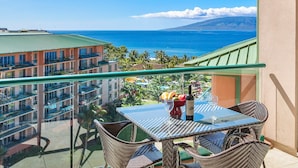 A balcony with a glass table and wicker chairs overlooking a coastal view with buildings and a mountain. - Maui Resort Rentals:  Honua Kai Konea 933 – 2 BR Corner w/ Sweeping 9th Floor Ocean and Black Rock Views! (Lahaina)