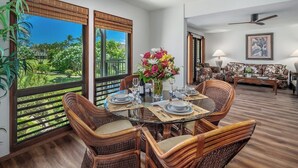 A dining area with wicker chairs, a glass table, and a view of a tropical garden through large windows. - Kahala 422 Poipu Tropical Nature By Suite Paradise (Koloa)