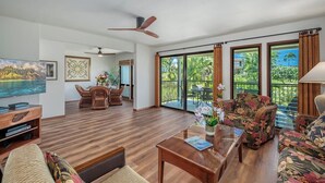 A spacious living area with a wooden floor, a ceiling fan, a television, and a dining area with chairs and a table. - Kahala 422 Poipu Tropical Nature By Suite Paradise (Koloa)