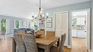 A dining area with a wooden table and wicker chairs, a chandelier, and a view of the kitchen and living room through an open door. - 47 S. Beach Ln | 4 Bedroom 5th Row Ocean | Pet Friendly | STR #029802 (Hilton Head Island)
