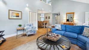 A spacious living room with a blue sofa, a round coffee table, and a dining area with a chandelier. - 47 S. Beach Ln | 4 Bedroom 5th Row Ocean | Pet Friendly | STR #029802 (Hilton Head Island)