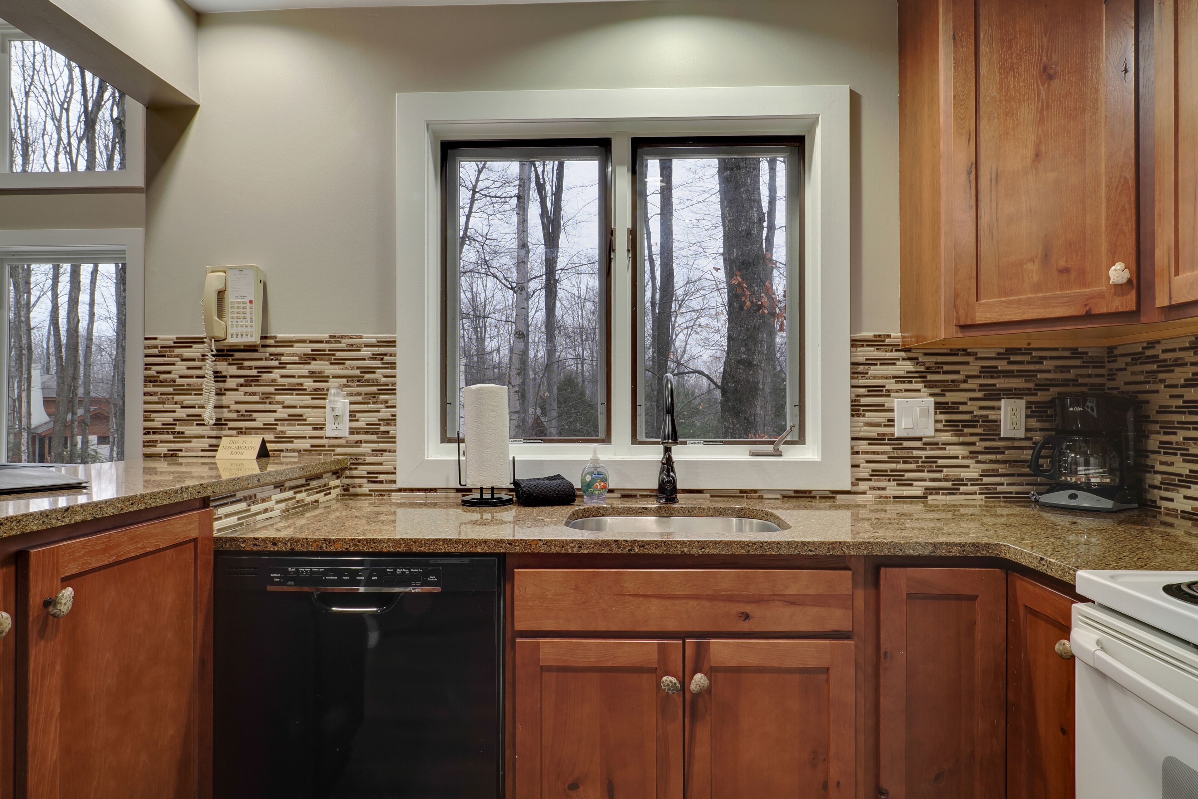 A kitchen with wooden cabinets, a dishwasher, a double sink, and a window with a view of trees.